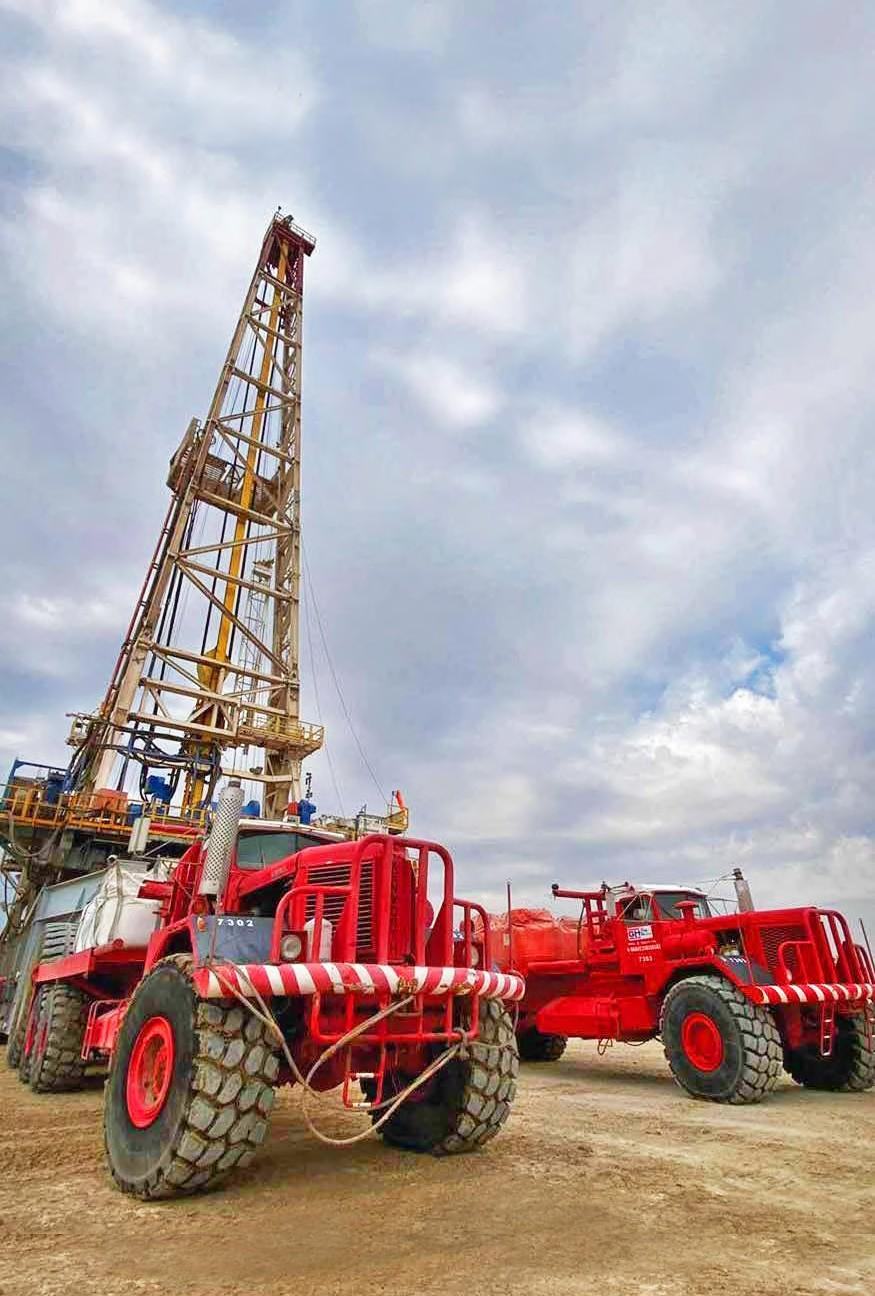 Red GH Rig Move oilfield trucks parked in front of a drilling rig mast under a cloudy sky